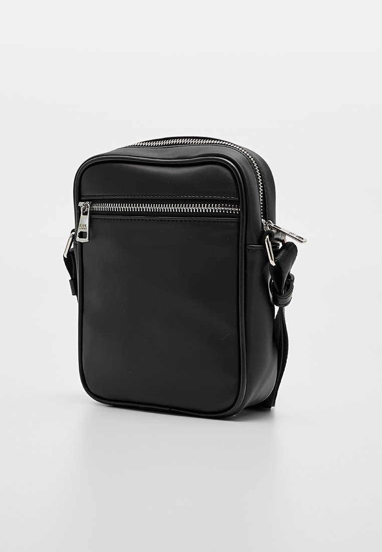 Bolso de cuero negro con forma rectangular, con un bolsillo frontal con cremallera, herrajes plateados y una correa ajustable. Textura suave.