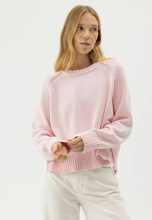 Jonge vrouw met lang blond haar, gekleed in een lichtroze gebreide trui en gebroken witte broek, kijkt iets naar links, handen bij de taille.