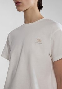 Valkoinen puuvillapaita lyhyillä hihoilla, jossa on pieni beige brodeerattu logo vasemmassa rintataskussa. Sileä pinta.