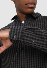 In primo piano, una persona indossa una camicia a maniche lunghe a scacchi neri e grigi con i polsini abbottonati, mentre sistema la manica.