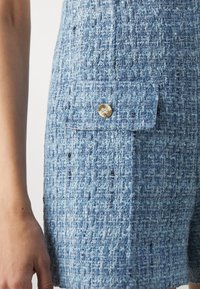 Gros plan d'une personne portant une jupe bleue texturée avec une poche à rabat fermée par un petit bouton rond doré sur le côté.