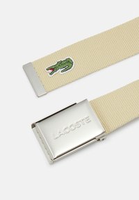 Béžový tkaný opasok so striebornou kovovou sponou s vygravírovaným nápisom "LACOSTE" a vyšitým zeleným dizajnom krokodíla.