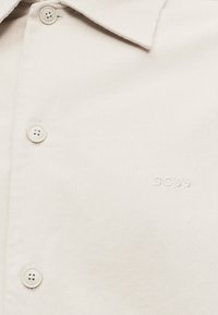 Chemise couleur crème avec boutons et col, avec "BOSS" brodé au niveau de la poche poitrine.