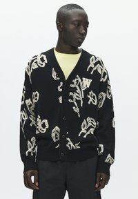 Crni kardigan s apstraktnim bež uzorcima, V-izrezom i kopčanjem na gumbe. Naglasak W logo na lijevoj strani. Model nosi svijetlo žutu majicu ispod.