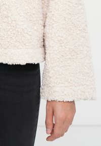 Main de personne reposant à côté d'un pantalon noir, portant une manche de veste en bouclés de couleur crème.