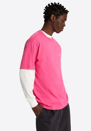 Jeune homme les mains dans les poches, portant une chemise rose vif à manches courtes sur une chemise blanche à manches longues et un pantalon sombre, de profil.