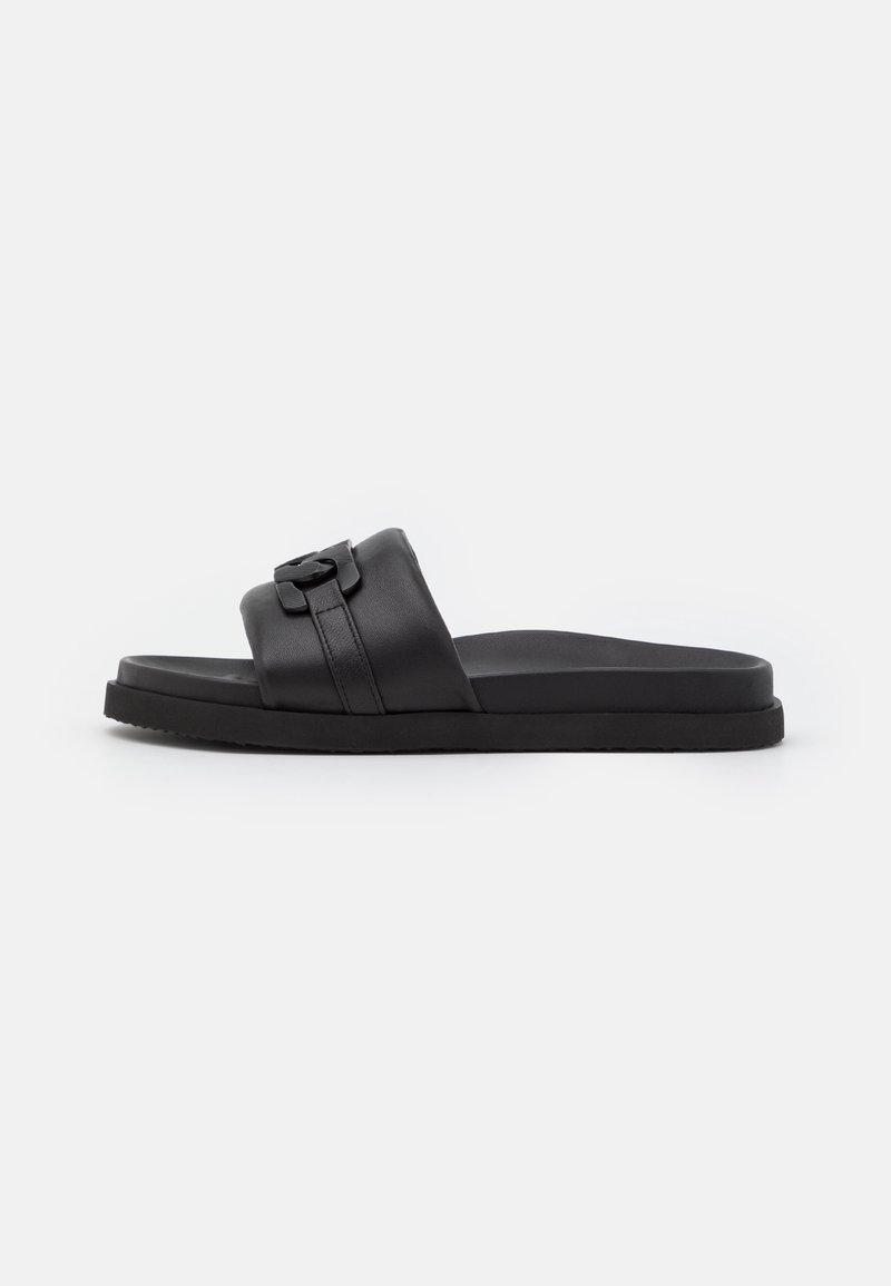 Sandale à glissière en cuir noir avec une large bride ornée d'un détail accentué, une semelle plate et une texture lisse. Design simple et minimaliste.