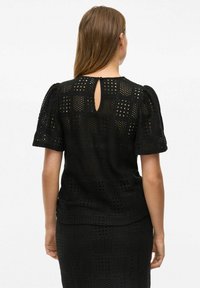 Haut noir à manches courtes avec un motif texturé en tissage ouvert et une fermeture à clé au dos. Le tissu semble léger et respirant.