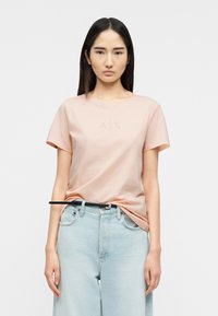 Νεαρή γυναίκα με μακριά σκούρα μαλλιά φοράει ανοιχτό ροζ μπλουζάκι A|X, το οποίο είναι μαζεμένο μέσα σε ανοιχτόχρωμο μπλε τζιν με μαύρη ζώνη, στέκεται μπροστά σε λευκό φόντο.