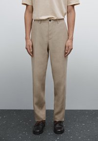 Beige Hose mit geradem Schnitt, aus leichtem Material. Mit einem einzelnen Knopfverschluss und seitlichen Taschen, kombiniert mit schwarzen Schuhen.