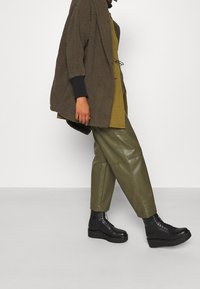 Cappotto khaki oversize in tessuto testurizzato, indossato su un top scuro, abbinato a pantaloni verde oliva a vita alta e stivali neri con tacco alto.