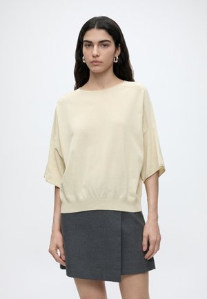 Kvinde med langt mørkt hår iført en løs beige top med korte ærmer og en mørkegrå nederdel mod en ensfarvet hvid baggrund.
