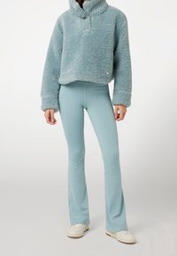 Měkký světle modrý svetr s vysokým límcem a knoflíkovým detailem, doplněný přiléhavými, rozšířenými světle modrými kalhotami a bílými teniskami.