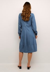 Gaiši zilā džinsa kleita ar garām piedurknēm, pieguļošu augšu un plandošu svārku. Iezīmēta ar šuvju akcentiem un pogām aizmugurē.