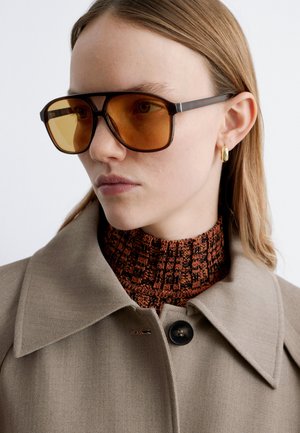 Femme portant de grandes lunettes de soleil teintées orange, une boucle d'oreille en or en forme d'anneau, un col roulé marron à motifs et un manteau beige à large col avec bouton.