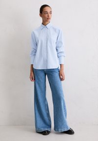 Light blue and white striped button-up shirt with a pointed collar, worn with loose blue jeans, featuring wide legs and a black shoe.
