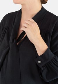 Schwarze Bluse mit einem gebundenen Kragen, gefalteten Schultern und geknöpften Manschetten. Weicher Stoff und leicht lockere Passform mit fließendem Design.