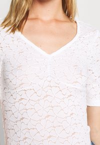 Bílý krajkový top s výstřihem do V, zdobený květinovými vzory a krátkými rukávy, s lehkou texturou a jemným designem.
