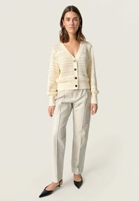 Krämfärgad stickad cardigan med v-ringning, öppet vävmönster och svarta knappar, kombinerad med ljusbeige läderbyxor och svarta klackar.