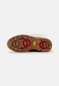 Suela de goma marrón con patrones de tracción profundos, con acentos de logotipo rojo y un detalle central amarillo. Borde superior suave de color crema.