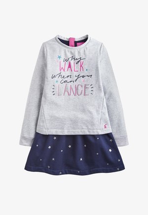 Sweat-shirt gris clair avec texte et accents scintillants, porté au-dessus d'une jupe bleu marine ornée de motifs d'étoiles argentées dispersées. Manches longues, col rond.
