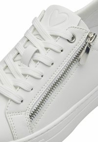Hvide sneakers lavet af glat syntetisk materiale med flade hvide snørebånd, en dekorativ lynlås på siden og et tekstureret hjerte-design på tungen.