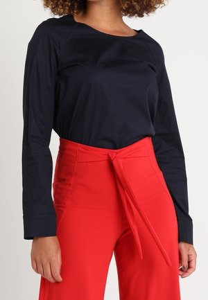 Kvinde iført en sort bluse med lange ærmer, stoppet ned i højtaljede røde bukser med et bindebånd foran, poserer foran en ensfarvet baggrund.