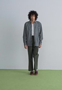 Geruite blouse in grijs en zwart over een wit T-shirt, gecombineerd met groene broeken en bordeauxrode schoenen. Krullend haar, staande op een groene vloer.