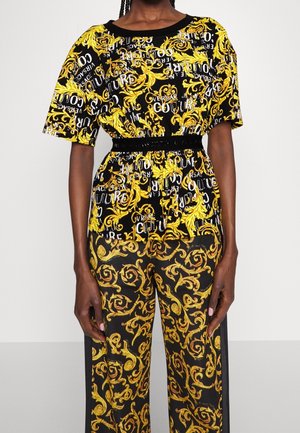 Czarno-żółty strój z barokowym wzorem, krótkimi rękawami, elastyczną talią i powtarzającym się napisem „Versace Couture” na górze.