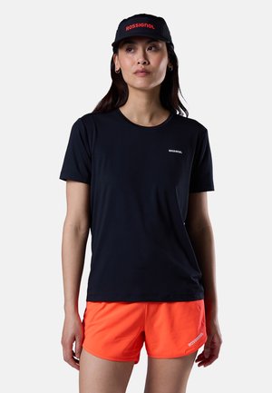 Femme portant une casquette noire Rossignol et un t-shirt avec un short Rossignol orange vif, debout devant un fond clair uni.