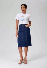 Denim midi skirt with front seam, side pockets, and contrasting orange stitching, paired with a white graphic t-shirt and white sandals.