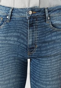 Detailný záber modrých džínsov s opakujúcim sa vzorom textu "JOOP! JEANS" a viditeľnými prednými vreckami, pútkami na opasok, zipsom a gombíkom.