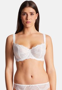 Soutien-gorge en dentelle blanche avec un motif floral, avec armatures de soutien et bretelles réglables. Bords festonnés notables et un petit nœud au centre.