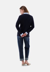 Marineblauer gerippter Pullover über einem hellblauen Kragenhemd, kombiniert mit dunkelblauen, hochgekrempelten Jeans und hautfarbenen High Heels.