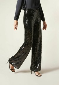 Pantalons large en sequins noirs avec une finition brillante et une surface texturée, associés à des talons noirs à lanières.