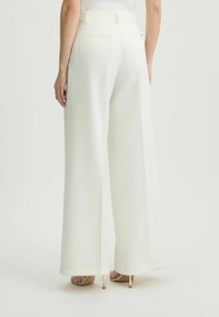 Wijd uitlopende broek in witte stof, met achterzakken en een gladde textuur. Gecombineerd met doorzichtige hoge hakken sandalen.