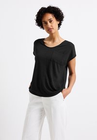 Haut noir à manches courtes avec décolleté rond et poche avant, fabriqué en tissu doux. Associé à un pantalon large de couleur claire.