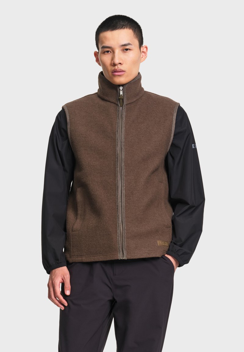 Homme portant une veste sans manches marron zippée par-dessus une chemise noire à manches longues, debout avec une main dans la poche devant un fond uni.