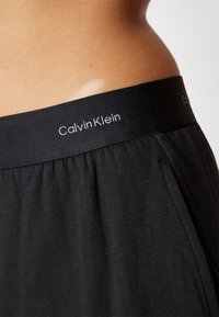 Zwarte elastische tailleband met "Calvin Klein" logo, gecombineerd met zacht, vloeiend zwart materiaal. Eenvoudig ontwerp met plooien voor extra textuur.