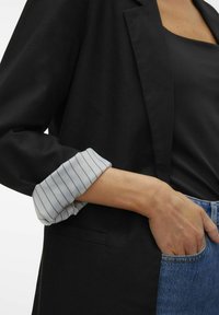 Blazer noir au design épuré, avec une manchette rayée blanche et grise. Gros plan sur la poche et le bras reposant contre un denim bleu.