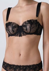 Reggiseno in pizzo nero con ferretto, motivo floreale, spalline in satin regolabili e dettagli in oro. Materiale trasparente con una fascia liscia.