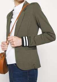 Blazer verde oliva con un design aderente, chiusura con un solo bottone e accenti a righe nere e bianche ai polsini. Abbinato a una maglietta bianca e jeans.