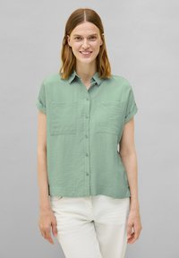 Camicia a maniche corte, di colore verde chiaro, con bottoni e due tasche frontali, realizzata in un tessuto testurizzato e con una vestibilità rilassata. I pantaloni bianchi completano il look.