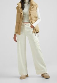 Beżowa kamizelka puchowa nałożona na białą, prążkowaną bluzkę, w połączeniu z szerokimi, białymi spodniami cargo i beżowymi butami na platformie. Widoczne fakturowane materiały i kieszenie.