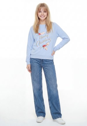 Világoskék pulóver "Boldog Karácsonyt" grafikával, rajta magyal levelek és bogyók. Széles szárú kék farmerrel és fehér tornacipővel párosítva.