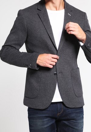 Mężczyzna zapinający ciemnoszary marynarkę na jeden guzik, na białej koszuli, w ciemnoniebieskich dżinsach, na jednolitym jasnym tle.