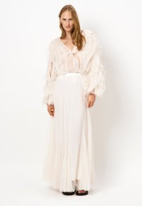 Lichte, doorschijnende witte top met geplooide ruffles. Lange, vloeiende witte rok met een aansluitende tailleband. Simpele zwarte sandalen complementeren de outfit.