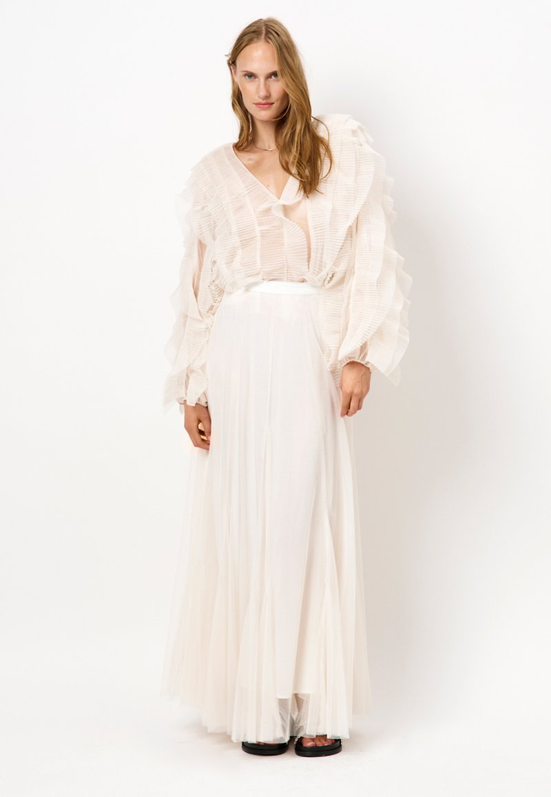 Lichte, doorschijnende witte top met geplooide ruffles. Lange, vloeiende witte rok met een aansluitende tailleband. Simpele zwarte sandalen complementeren de outfit.