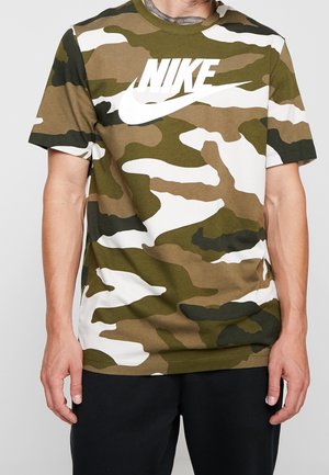 Man wearing green, brown, black, and white camo Nike t-shirt with large white logo, paired with black pants, hands relaxed at sides.