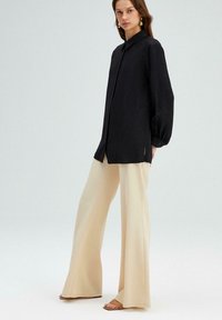 Vrouw in een losse zwarte lange mouw shirt met beige wijde broek en tan open teen sandalen, staande tegen een effen lichte achtergrond.